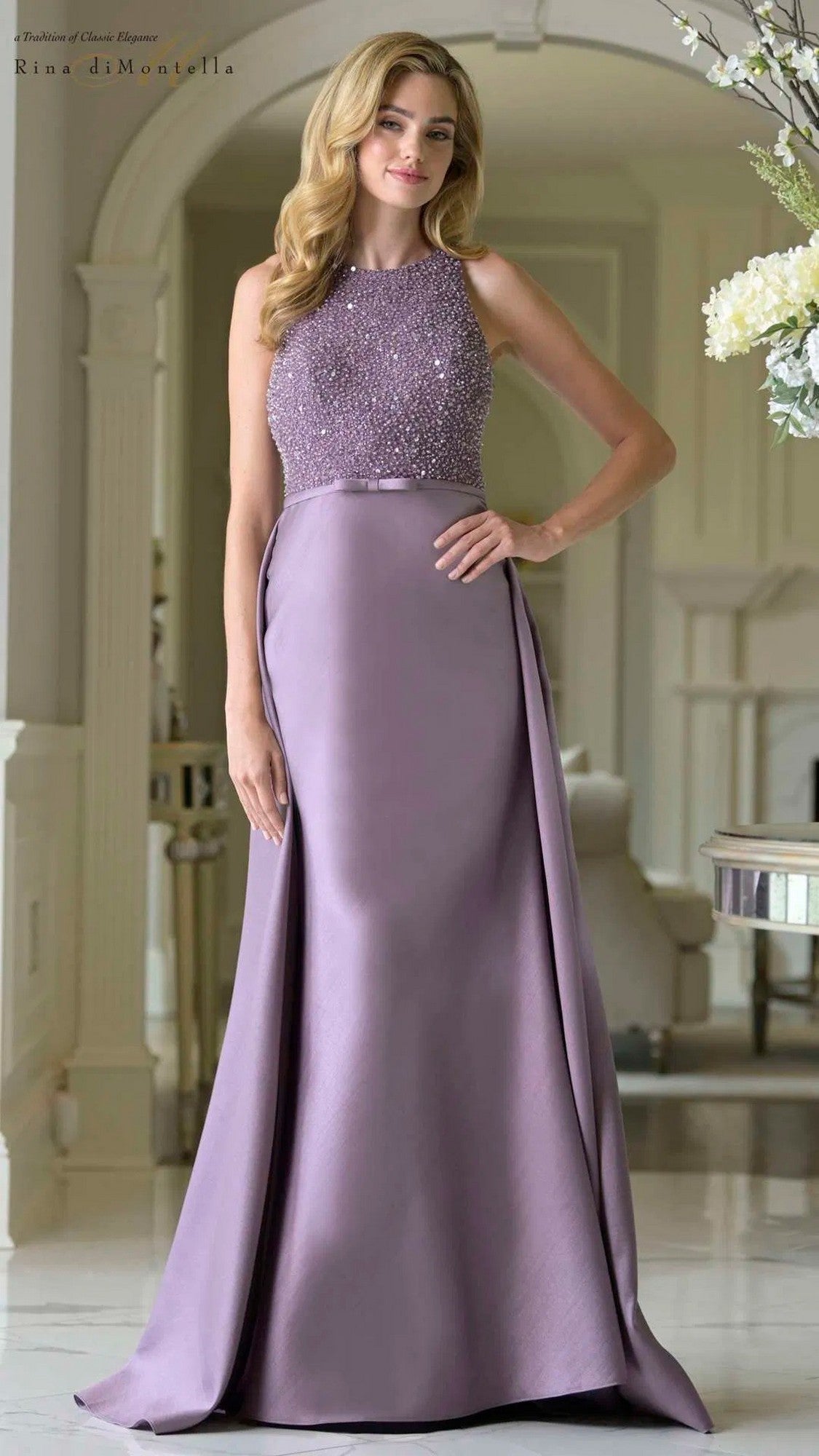 A woman stands indoors wearing the Long Formal Dress: Rina Di Montella RD3404, which features a beaded bodice and a flowing, floor-length satin skirt in soft lavender.